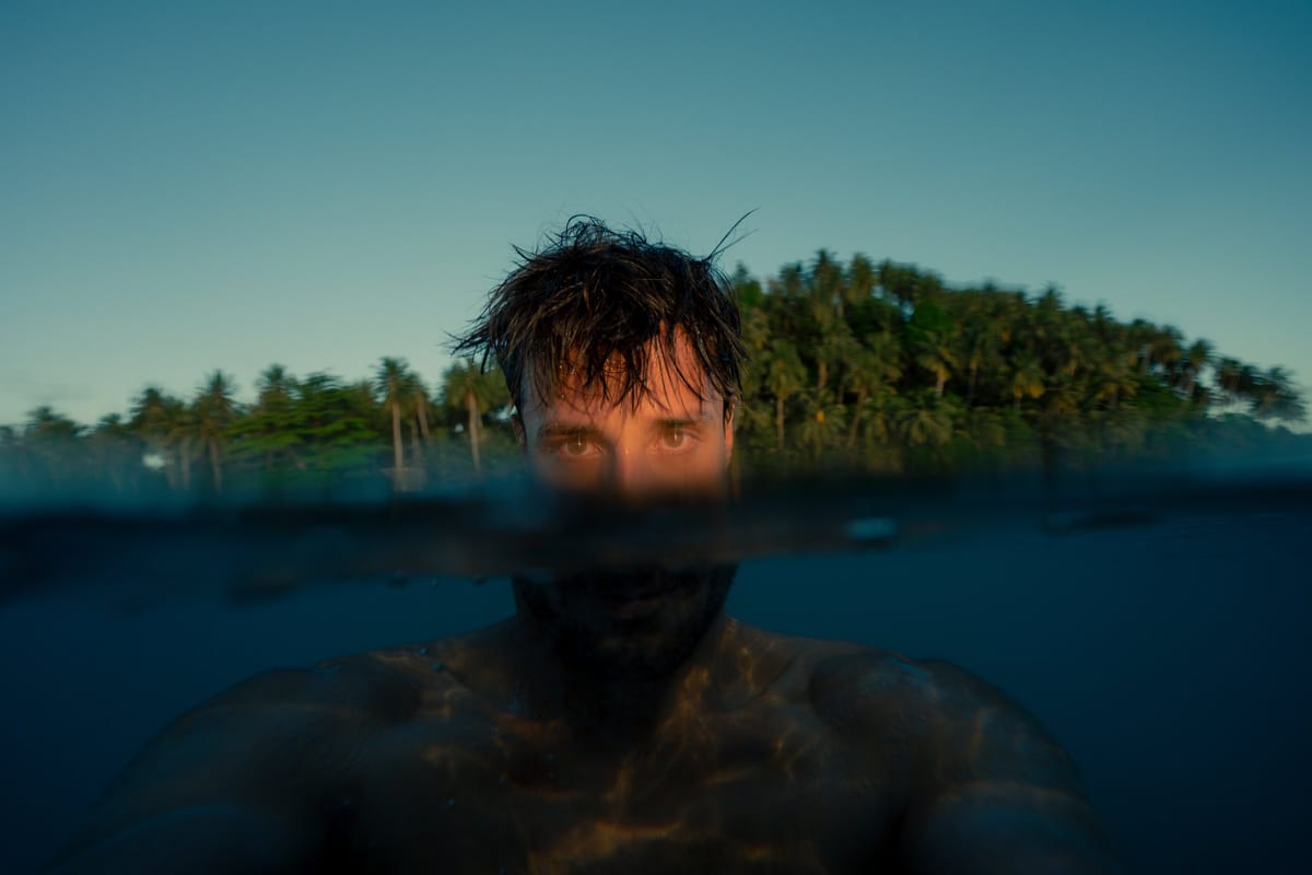 Adrien Desnoyer in the water, face half submerged, a palm-covered island behind him